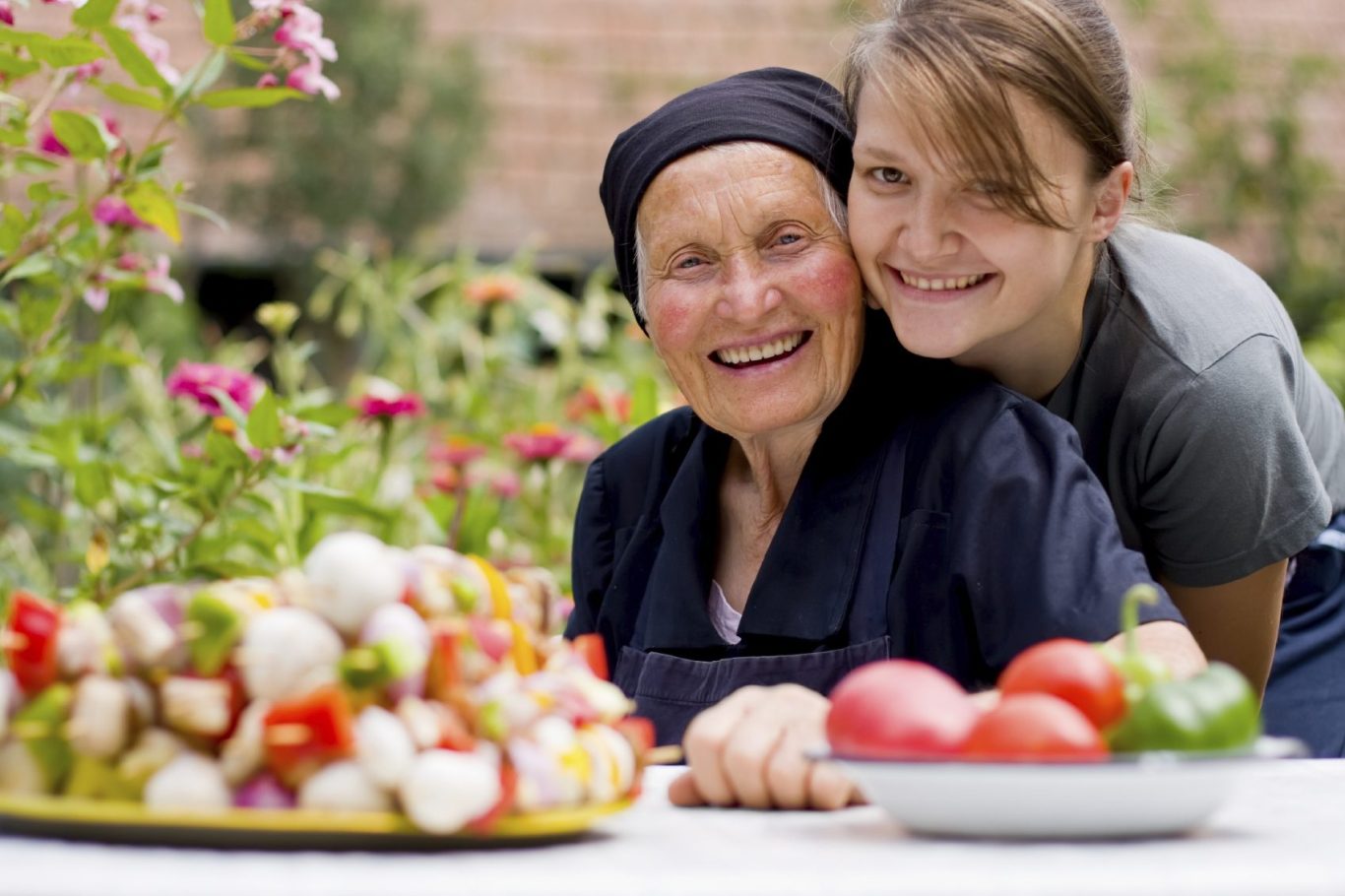 Two women smiling in a garden, with a platter of fresh vegetables in front of them.